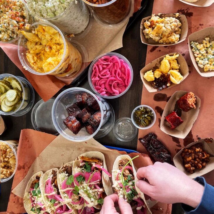 A table filled with tacos, pickled onions, baked beans, coleslaw, chips, cornbread, burnt ends, and sides—just what you'd expect from the best BBQ Charlotte NC has to offer. Two hands are assembling tacos topped with veggies and pickled onions.