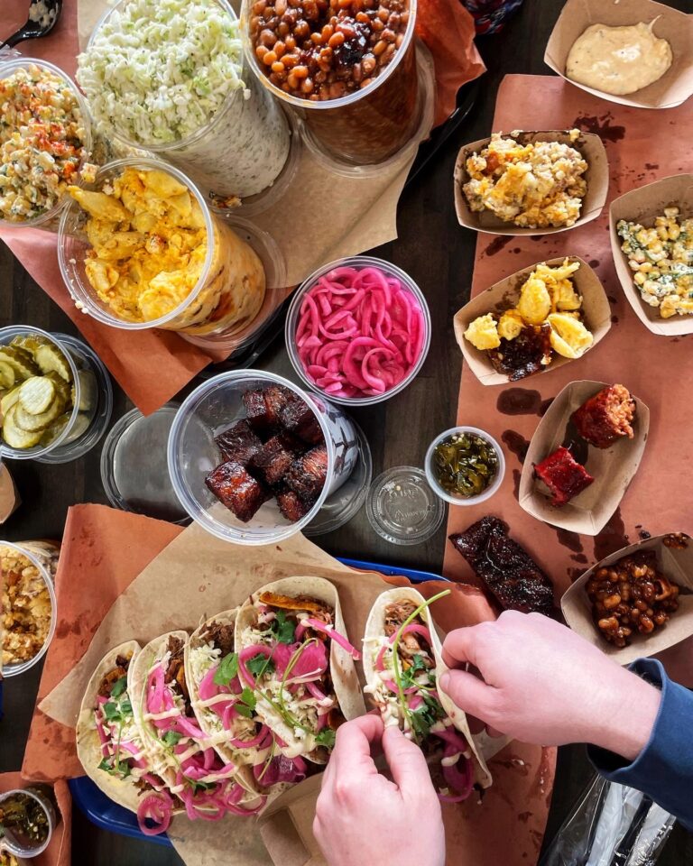 A table filled with tacos, pickled onions, baked beans, coleslaw, chips, cornbread, burnt ends, and sides—just what you'd expect from the best BBQ Charlotte NC has to offer. Two hands are assembling tacos topped with veggies and pickled onions.