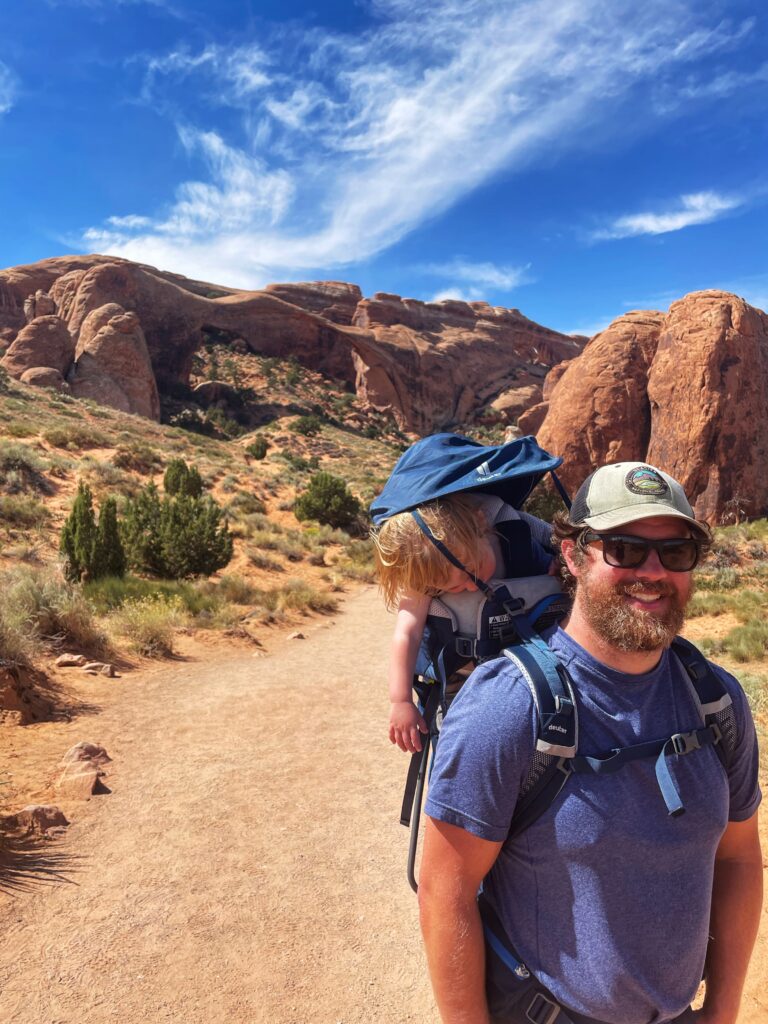 dad with son in a hiking backpack taking a nap with landscape arch in the background