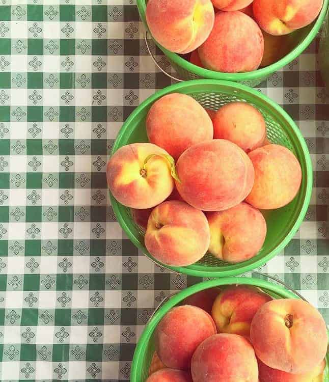 Three green baskets filled with ripe peaches sit on a green and white checkered tablecloth with a decorative pattern.