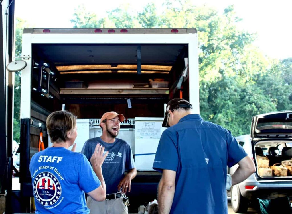 Three people stand and talk at the open back of a truck. One person wears a blue Staff shirt, another is laughing and wearing a cap, and a third person stands with their back to the camera. A car with the trunk open is nearby.
