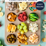 A muffin pan filled with kid-friendly snacks like fruit, cheese, pretzels, popcorn, and candy sits on a blue surface. Scattered snacks surround it. Text reads: “How to Make a Kids Charcuterie Board. Easy Recipe.”.