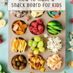A muffin tin filled with snacks—grapes, cucumber, raspberries, pretzels, cheese, olives, popcorn, and more—sits on a blue surface. Scattered snacks surround it. Text overlay reads: “How To Make a Kids Charcuterie Board.”.