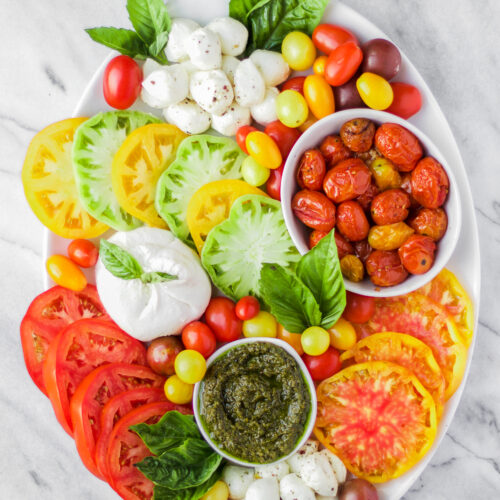 A platter with sliced red and yellow tomatoes, fresh mozzarella balls, burrata cheese, roasted cherry tomatoes, pesto, basil leaves, and colorful grape tomatoes, all arranged neatly on a white marble surface.