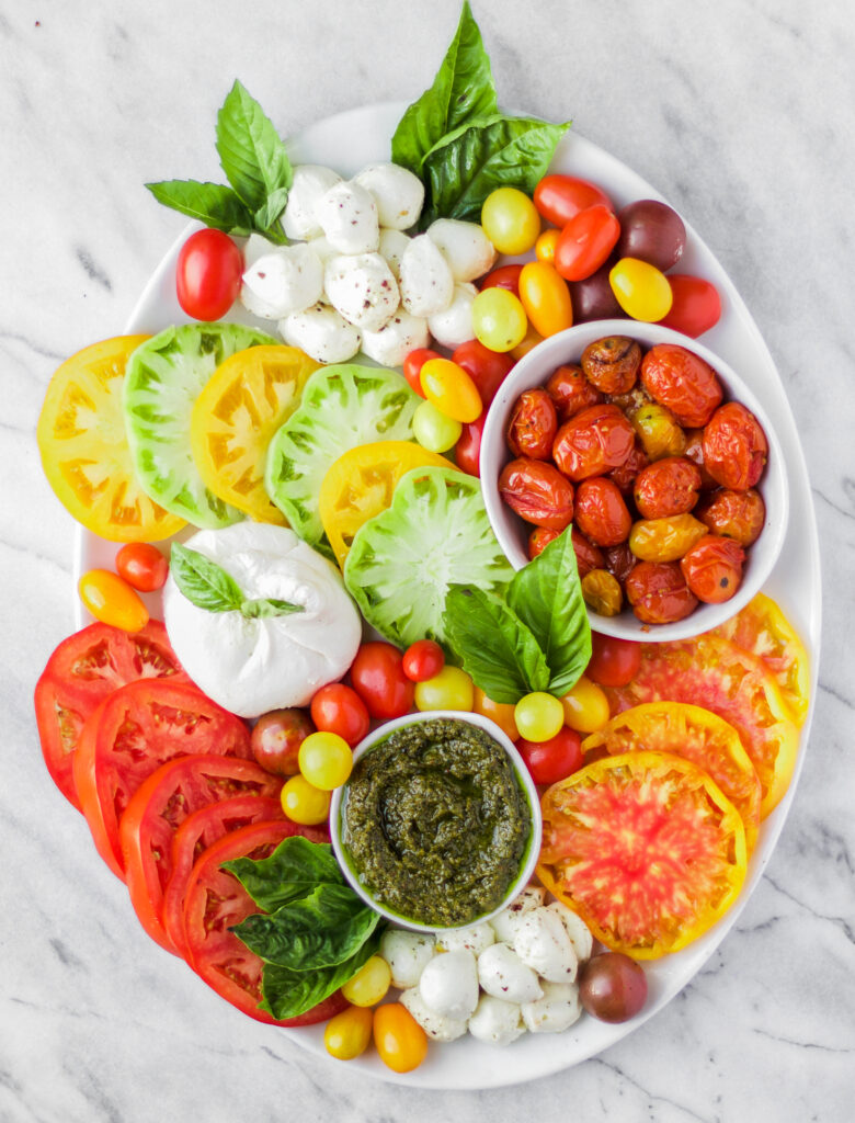 A platter with sliced red and yellow tomatoes, fresh mozzarella balls, burrata cheese, roasted cherry tomatoes, pesto, basil leaves, and colorful grape tomatoes, all arranged neatly on a white marble surface.