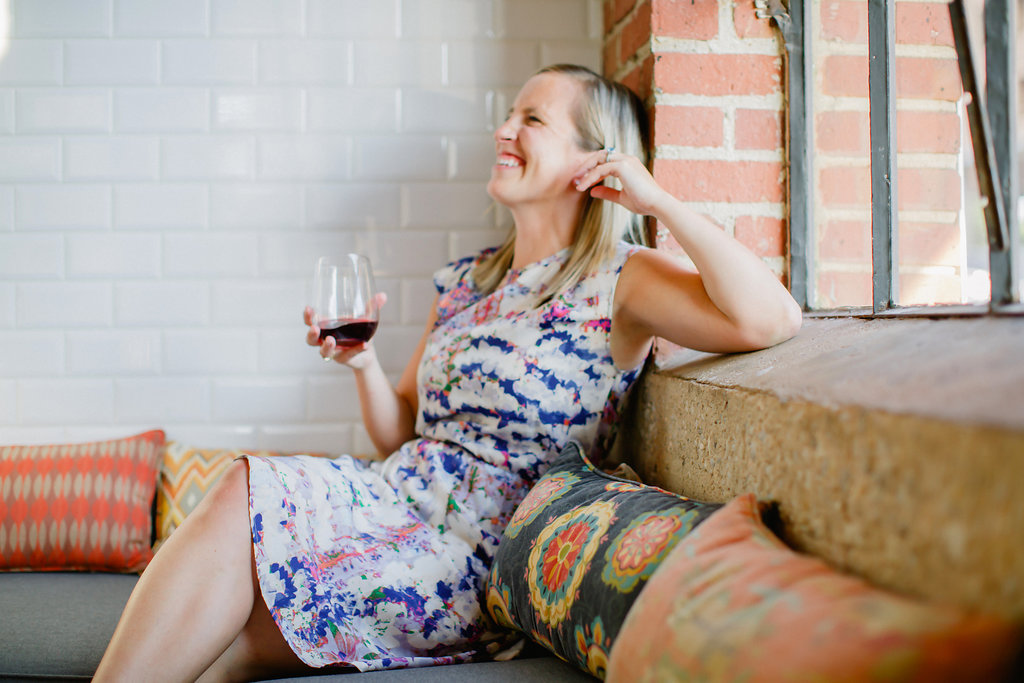 A woman in a floral dress sits on a cushioned bench by a window, smiling and holding a glass of red wine surrounded by colorful pillows.