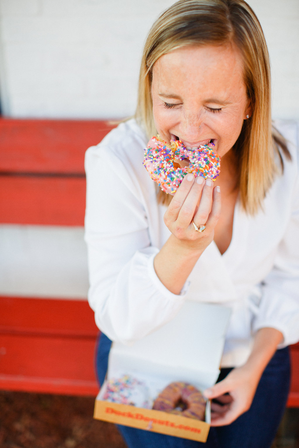 A woman with blonde hair and a white shirt sits on a red bench, smiling with her eyes closed as she takes a bite of a sprinkled donut. An open box of donuts rests on her lap.