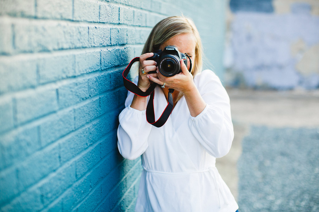 A person with blonde hair, wearing a white top, stands against a teal brick wall while holding a Canon camera up to their face.