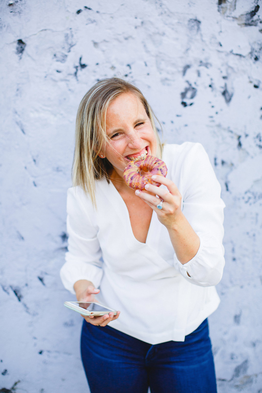 A woman in a white blouse and blue jeans smiles as she bites into a colorful donut, holding a smartphone in her other hand against a textured light gray wall.