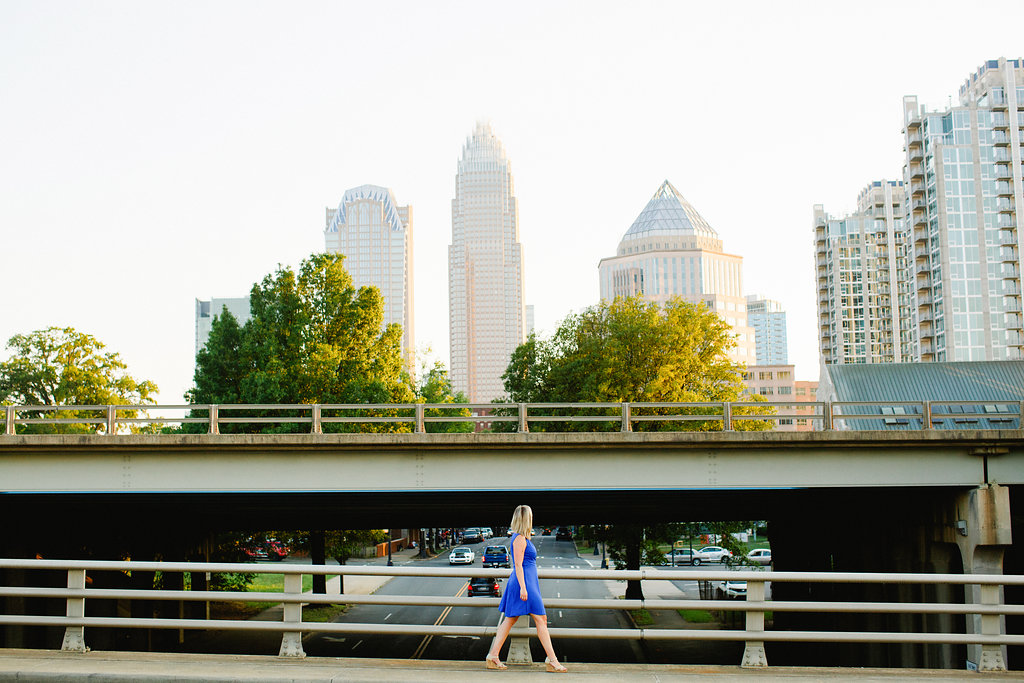 A woman in a blue dress walks across a bridge, with a city skyline and tall buildings in the background on a clear day, exploring an off the eaten path route through the city.