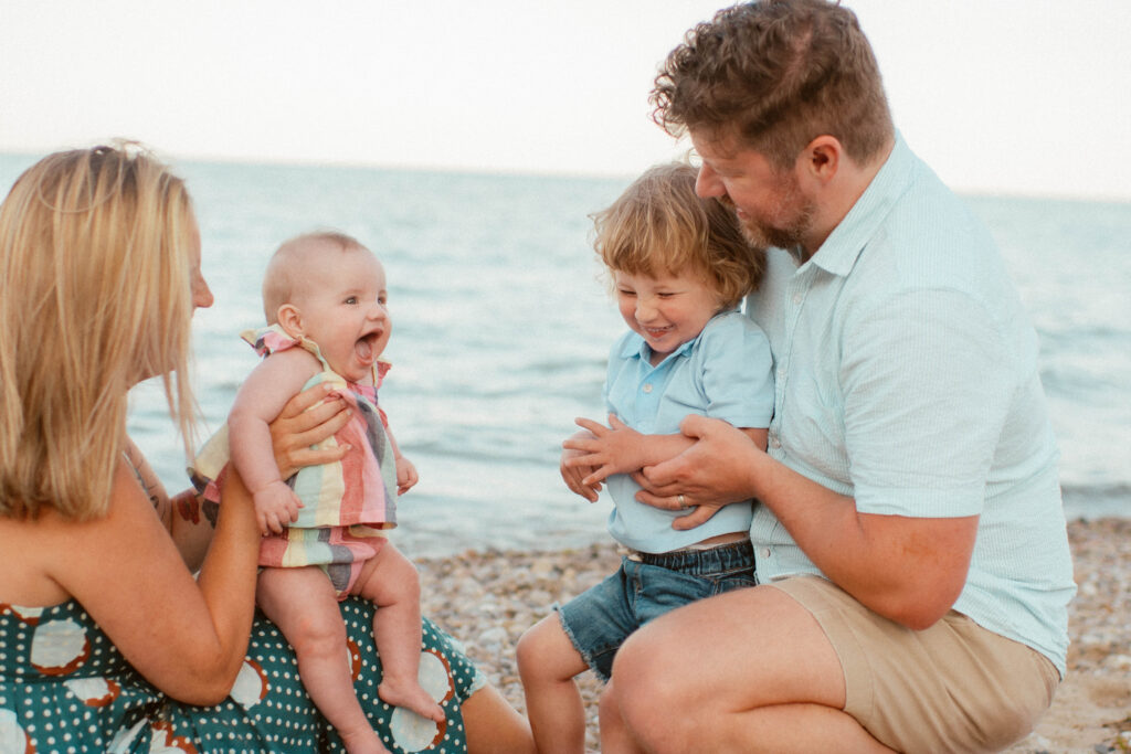 A family of four sits on a pebble beach by the water. The mother holds a smiling baby while the father and young child laugh, everyone happy and relaxed in this peaceful outdoor setting.