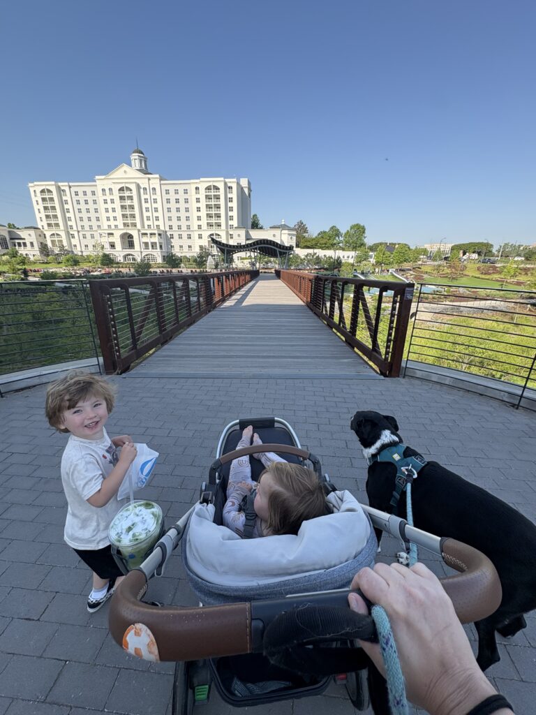 A person pushes a stroller with a baby, a smiling young child stands nearby holding snacks, and a black dog is on a leash. They are on a bridge with a large white building and greenery in the background under a clear sky.