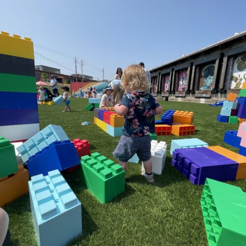 A toddler with blonde hair stands on grass among large colorful toy building blocks—one of many fun toddler activities Charlotte NC families enjoy—while other children and adults play in the background under a clear blue sky.