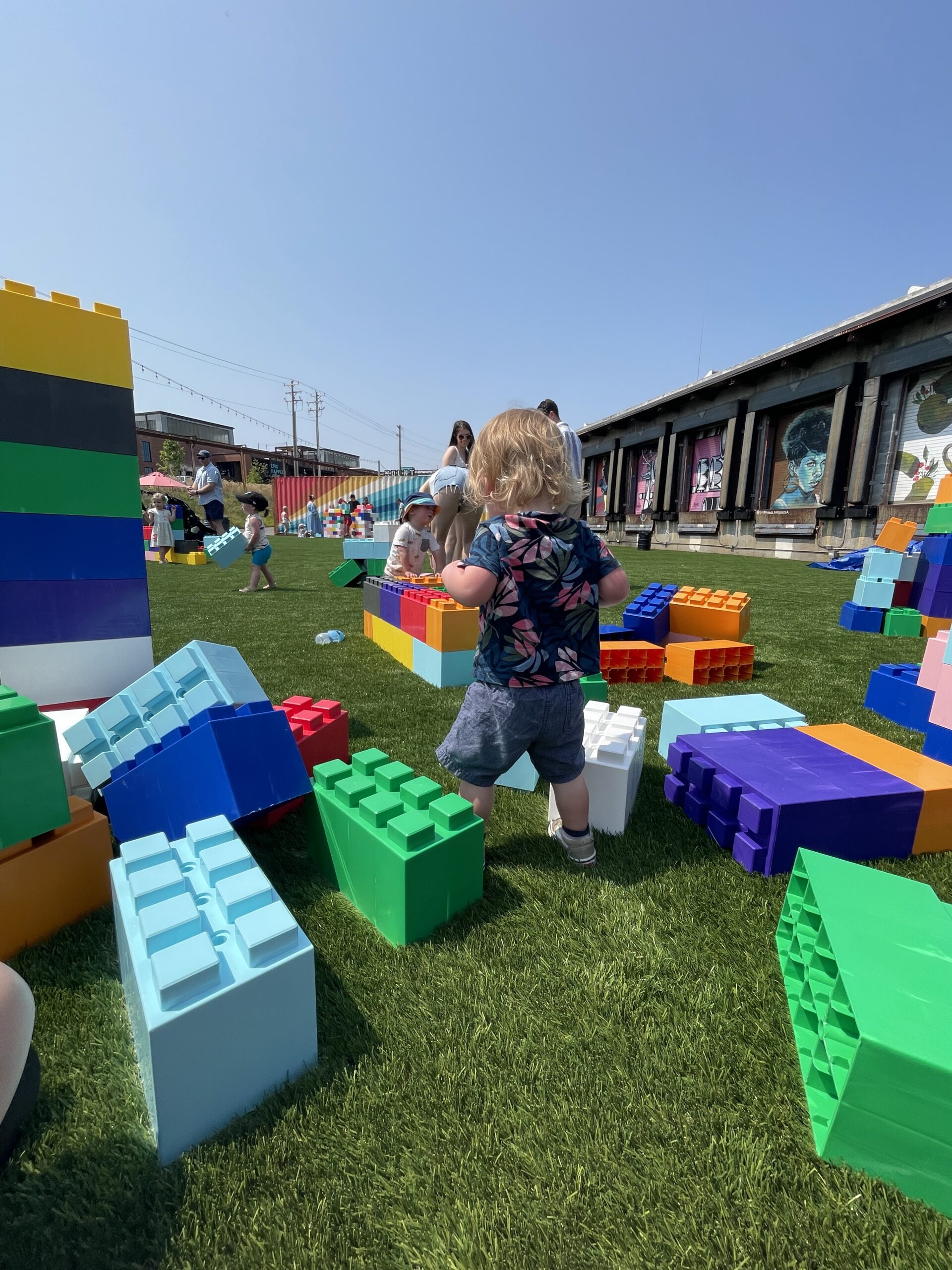 A young child with blonde hair walks on green grass among large, colorful toy blocks, with more children and adults playing in the background under a clear, sunny sky.
