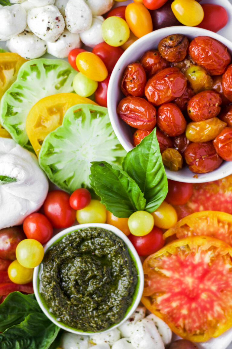 A colorful spread of fresh tomatoes in various colors, mozzarella balls, roasted tomatoes in a bowl, a bowl of green pesto, basil leaves, and slices of heirloom tomatoes arranged on a platter.