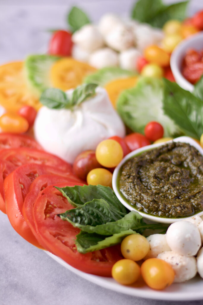 A platter with sliced tomatoes, yellow and red cherry tomatoes, fresh mozzarella, a ball of burrata cheese, green pesto in a small bowl, and fresh basil leaves arranged on a white surface.