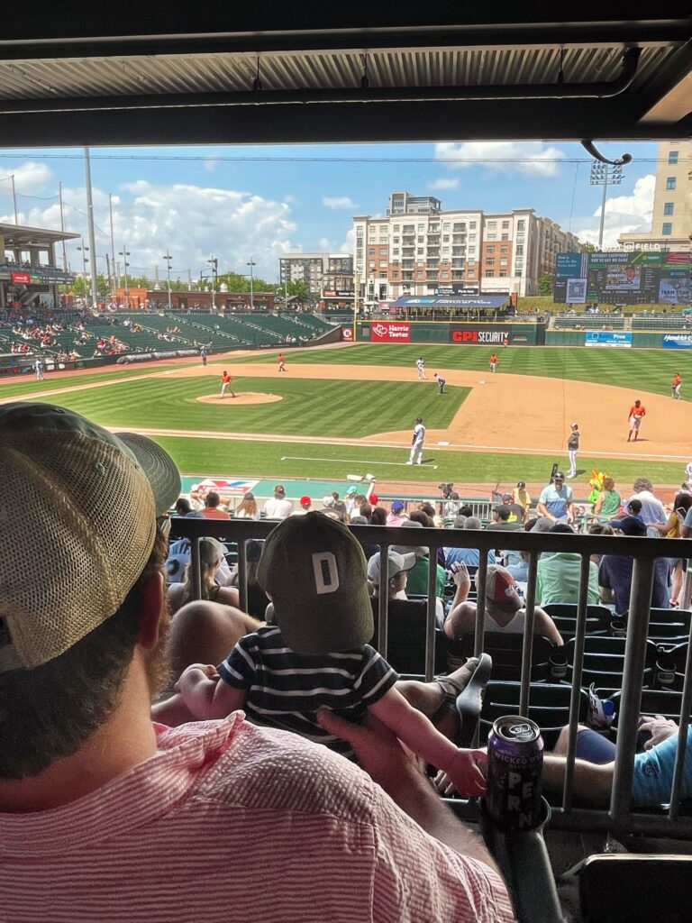 A man holding a baby overlooks a baseball game from stadium seats. The baby wears a cap with a D on it. The field, players, stands, and city buildings are visible under a partly cloudy sky.