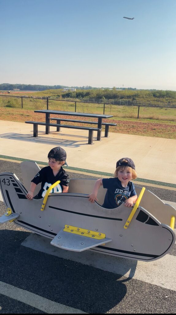 Two young children wearing baseball caps sit in a playground airplane ride-on toy. A picnic table and grassy field are in the background, with an airplane visible in the sky above the horizon.