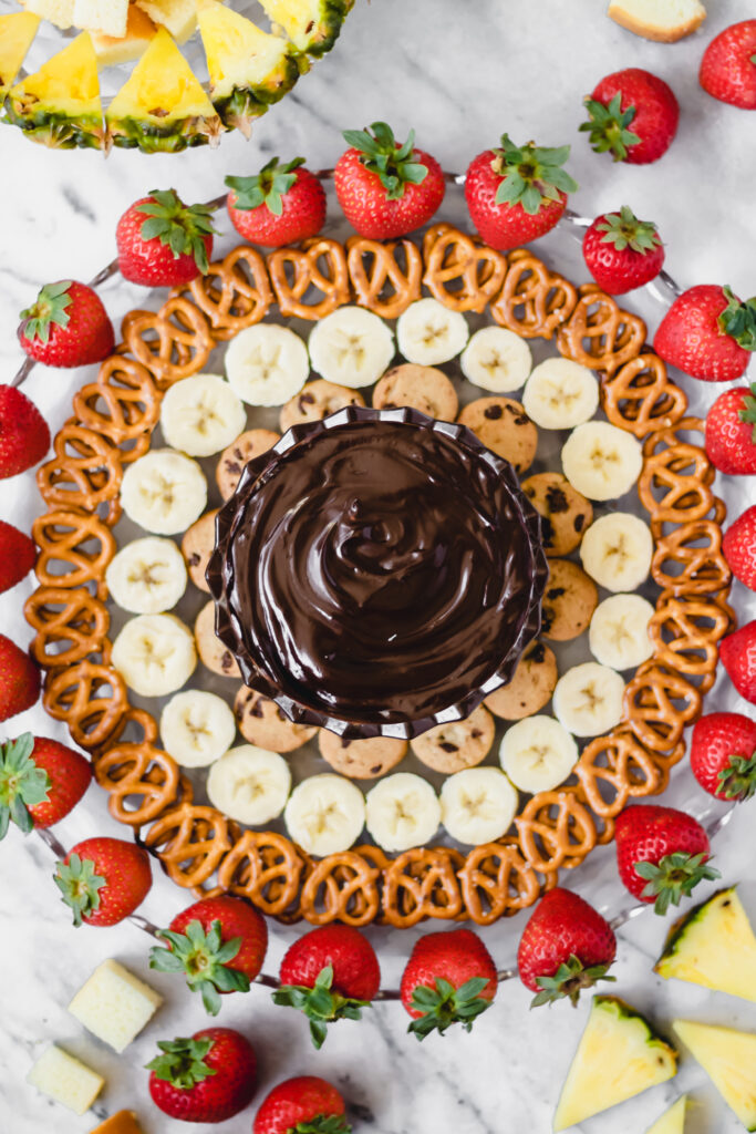 A circular platter arranged with concentric rings of strawberries, pretzels, banana slices, and cookies around a central bowl of chocolate dip, with pineapple pieces in the corners.