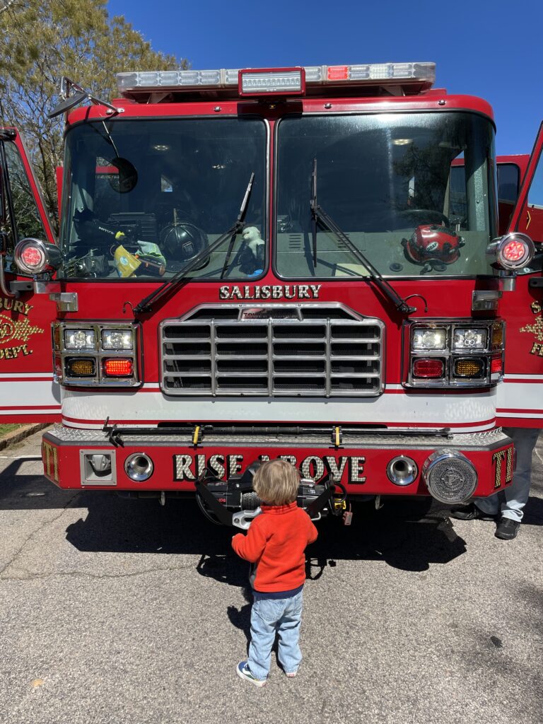 A young child in a red sweater stands in front of a large red Salisbury fire truck with open doors, looking up at its shiny grill and equipment on a sunny day.