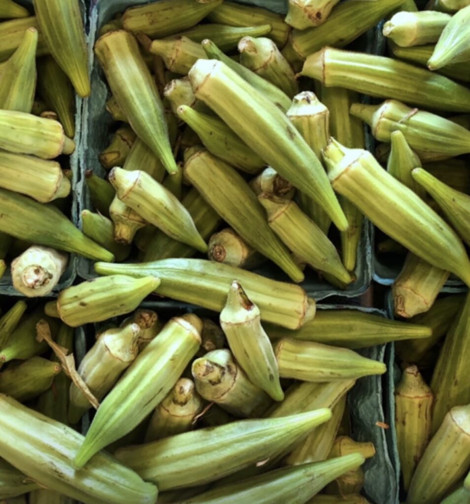 Close-up of several green okra pods piled in small cardboard cartons, displayed at what appears to be the Charlotte Regional Farmers Market.