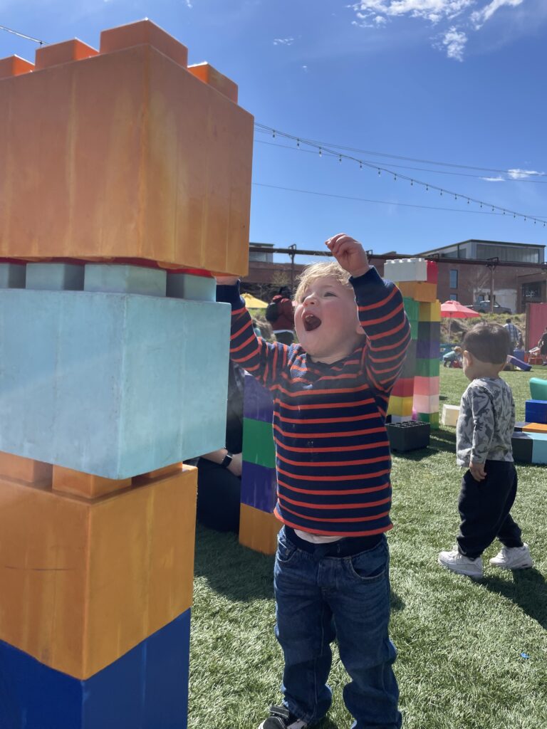 A young child smiles and reaches up toward giant colorful toy blocks outdoors on a sunny day, with another child and play structures visible in the background.