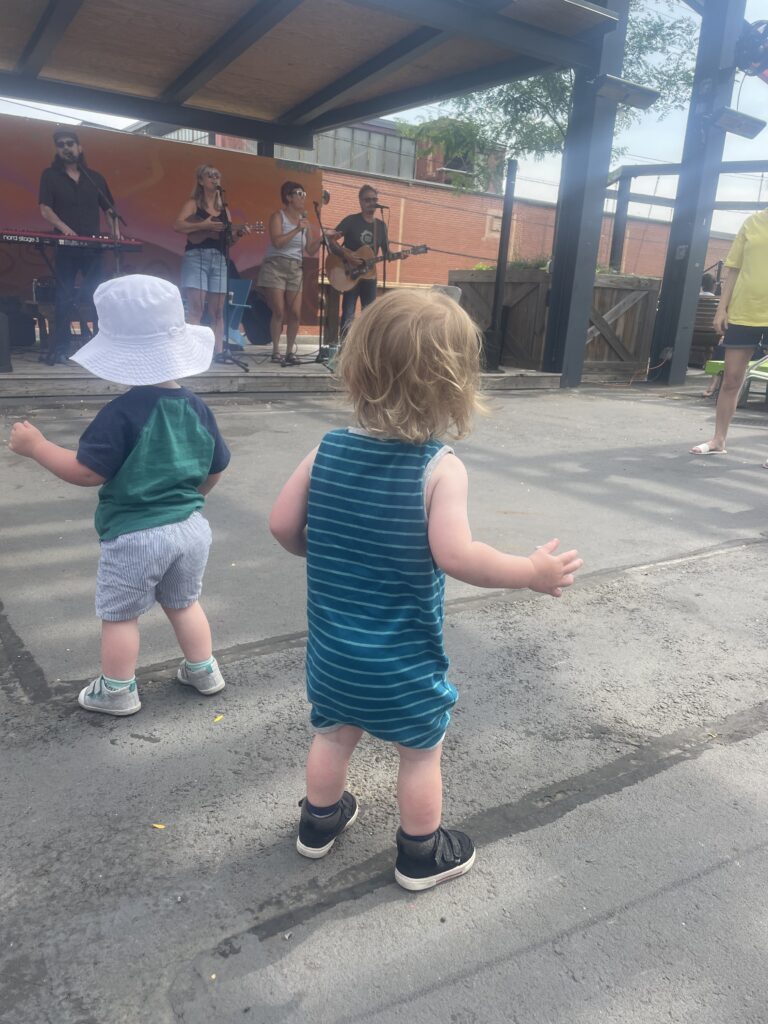 Two toddlers stand on a concrete surface, facing a stage where a band is performing outdoors. The toddlers appear to be dancing, and the scene is bright and sunny with people in the background.