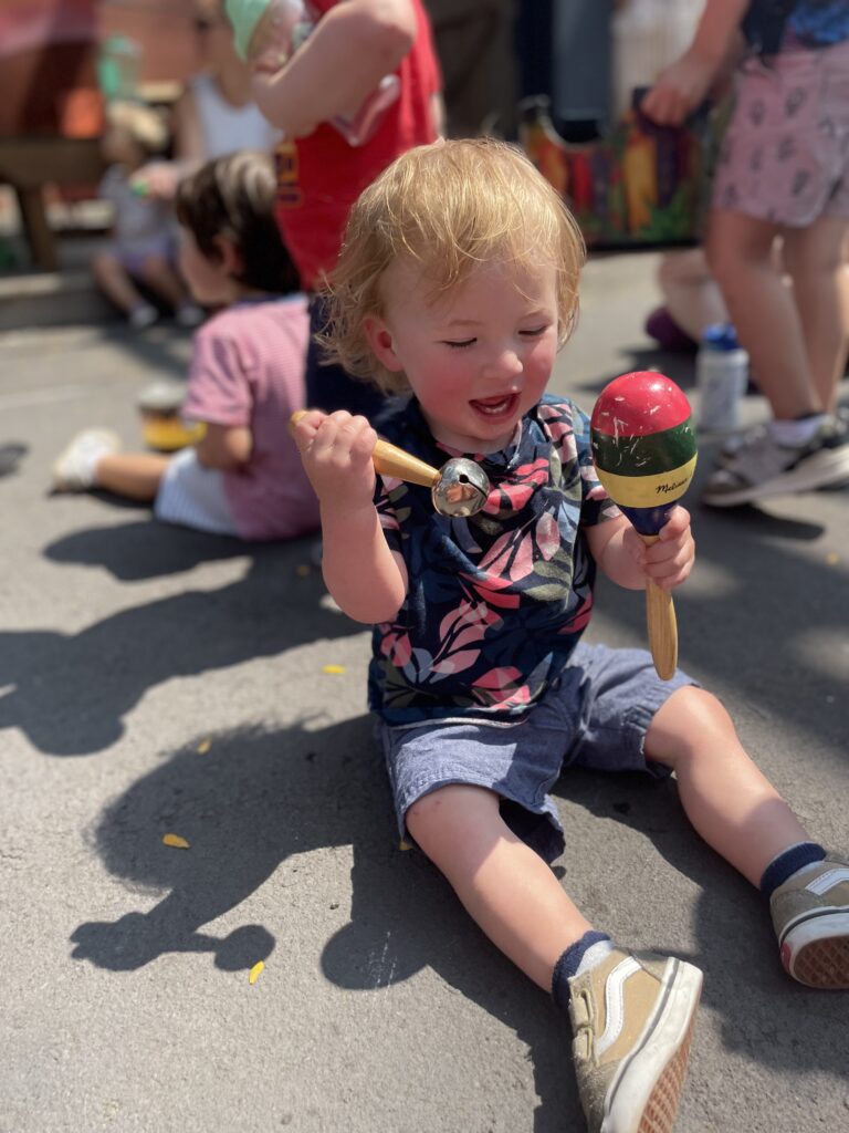 A toddler with blond hair sits on the ground outside, smiling and holding a maraca and a bell. Other children play in the background on a sunny day.