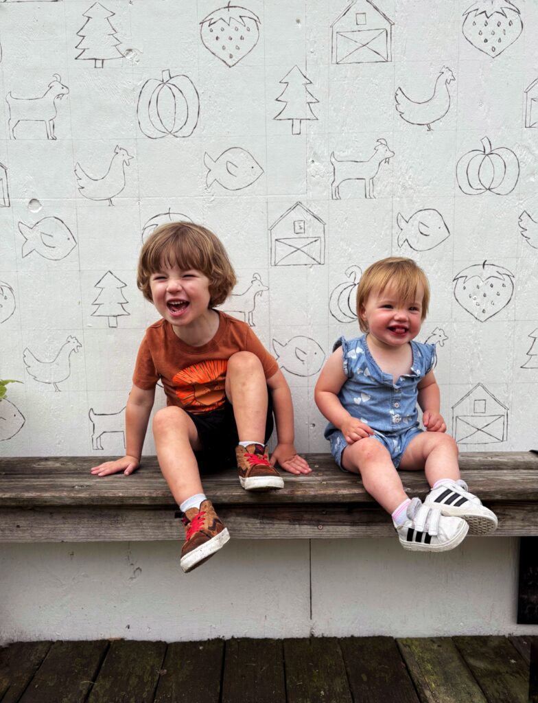 Two young children sit smiling on a wooden bench in front of a white wall decorated with simple line drawings of animals and farm items. The boy wears a brown shirt and shorts, and the girl wears a blue romper and white shoes.