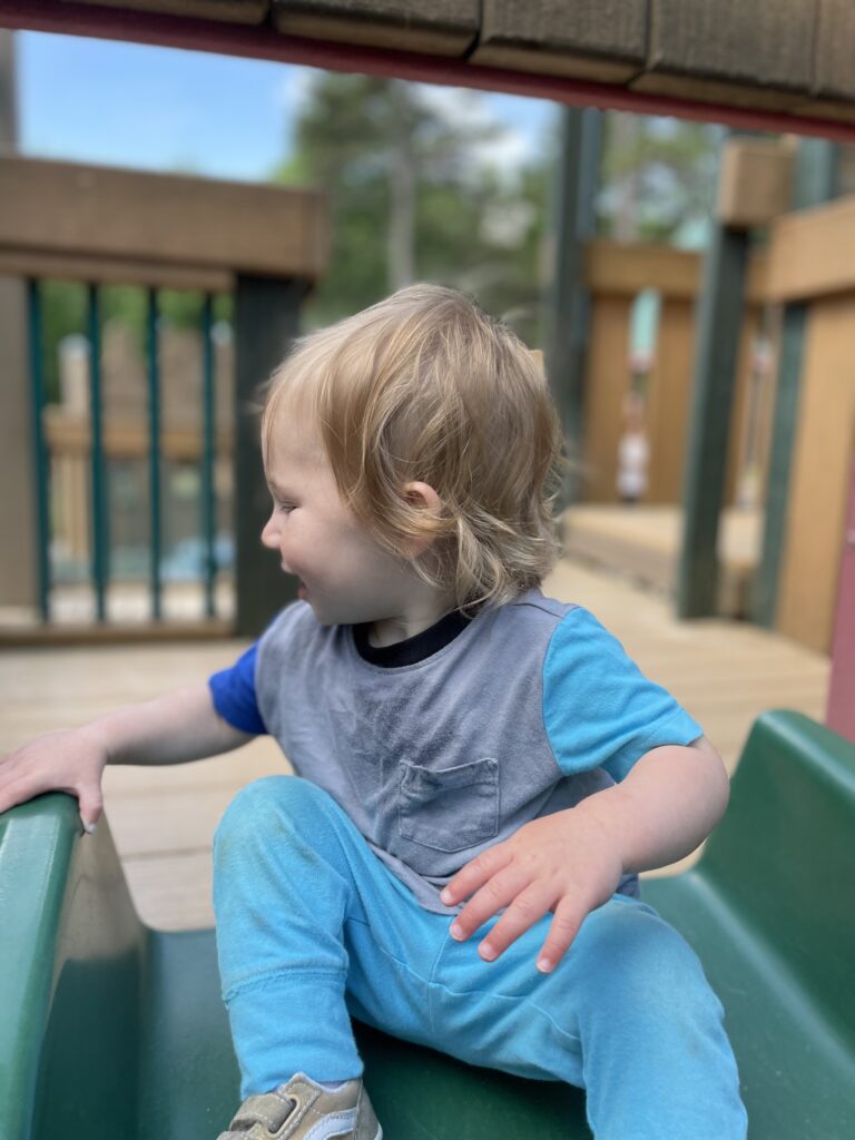 A young child with blond hair, wearing a blue and gray outfit, sits sideways on a green playground slide, looking to the left and smiling. The playground structure and trees are visible in the background.