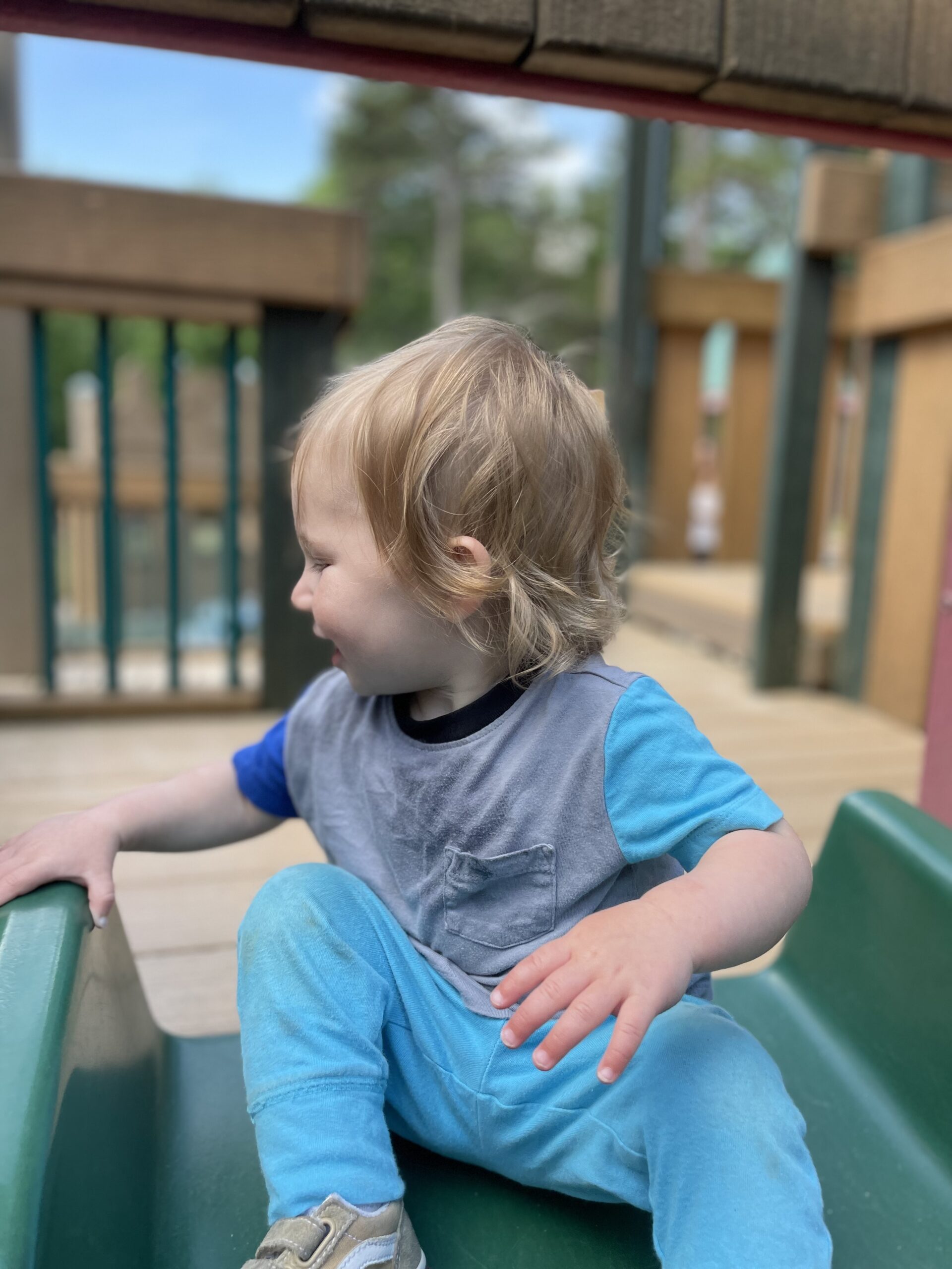 A young child with blonde hair, wearing a blue and gray outfit, sits sideways at the top of a green playground slide in Charlotte, NC—an example of fun toddler activities amid wooden playground structures and trees.