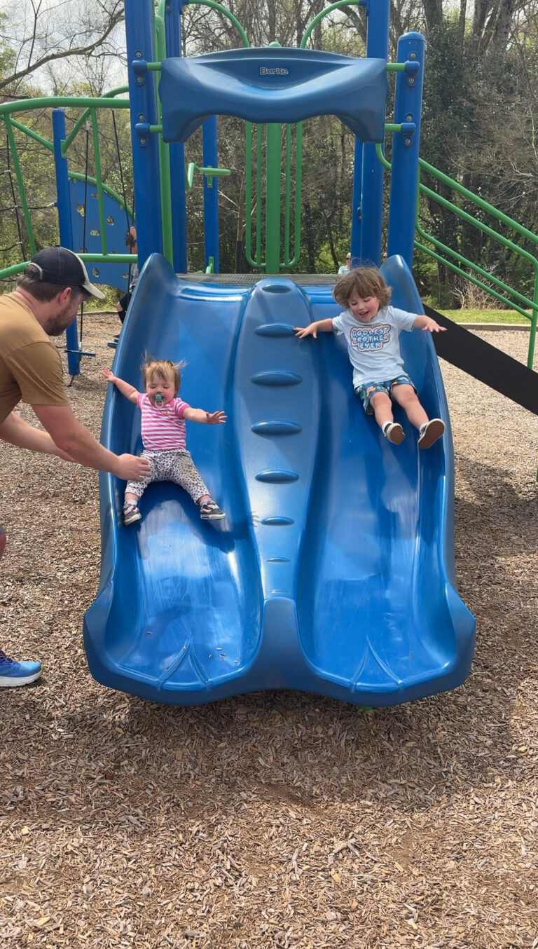 A man helps a young girl in a pink striped shirt as she slides down a blue double slide next to a boy in a blue shirt at a playground. Trees and playground equipment are visible in the background.