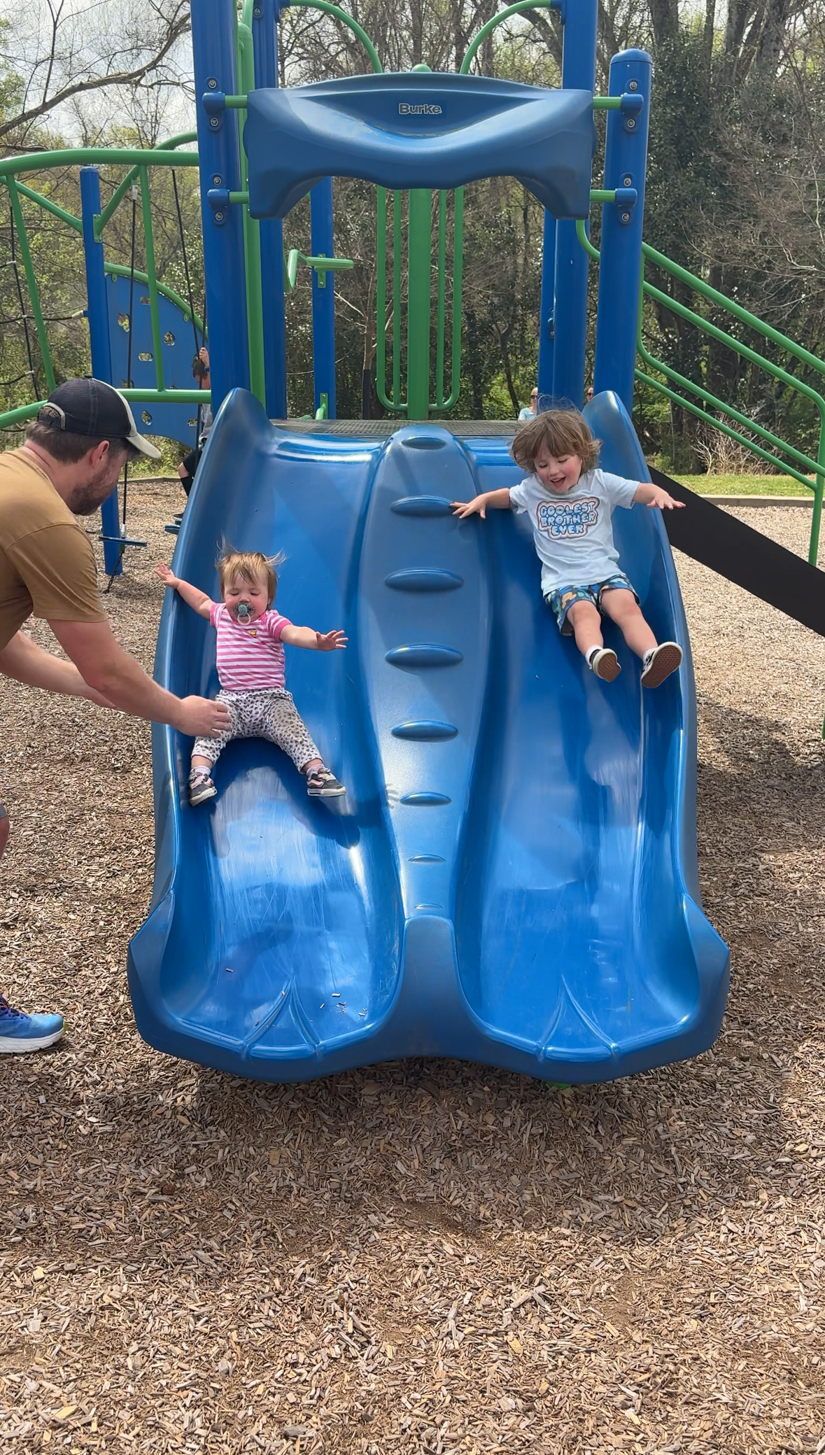 A man helps a young girl in a pink striped shirt as she slides down a blue double slide next to a boy in a blue shirt at a playground. Trees and playground equipment are visible in the background.