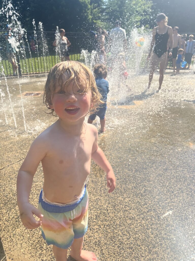 A young child in colorful shorts smiles while playing in a sunny splash pad with water fountains. Other children and adults are in the background, also enjoying the water on a bright day.
