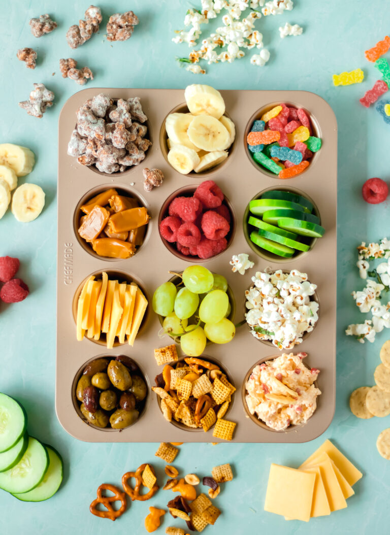 A muffin tin filled with various snacks including popcorn, banana slices, gummy worms, caramel candies, raspberries, cucumber slices, sliced cheese, green grapes, snack mix, pimento cheese, and olives. Snacks are scattered around it.