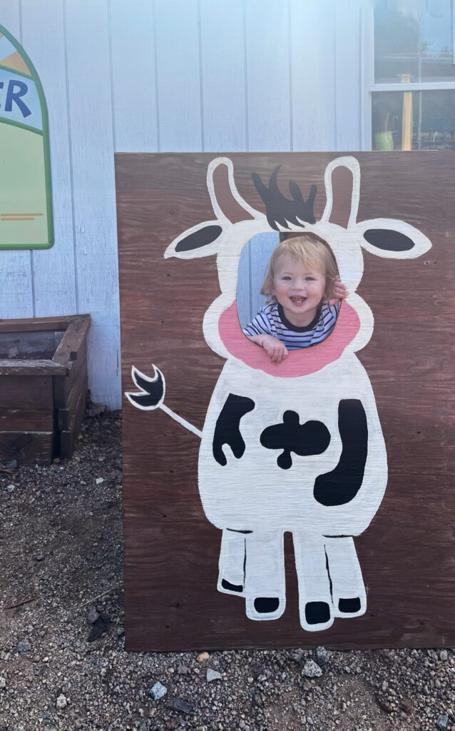 A smiling toddler poses with their head through a cutout of a cartoon cow on a wooden board outdoors, creating the illusion that they have the body of the cow.