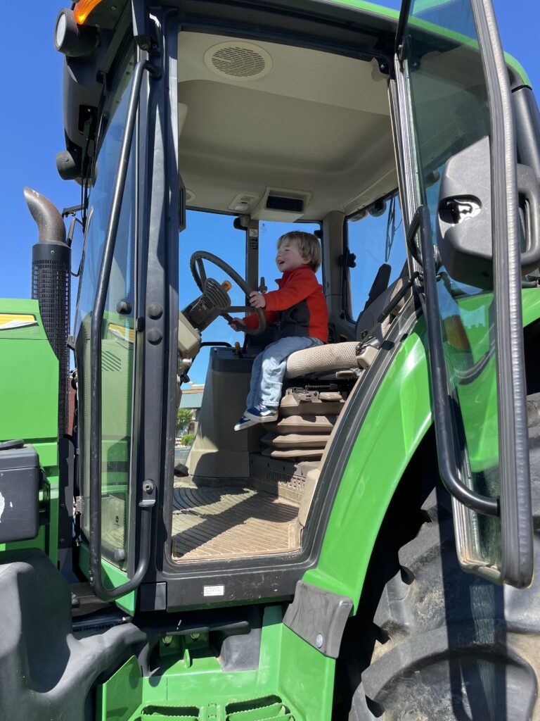 A young child in a red shirt sits in the driver’s seat of a large green tractor, holding the steering wheel and looking forward. Sunlight shines through the cab’s windows, and the sky is clear and blue outside.