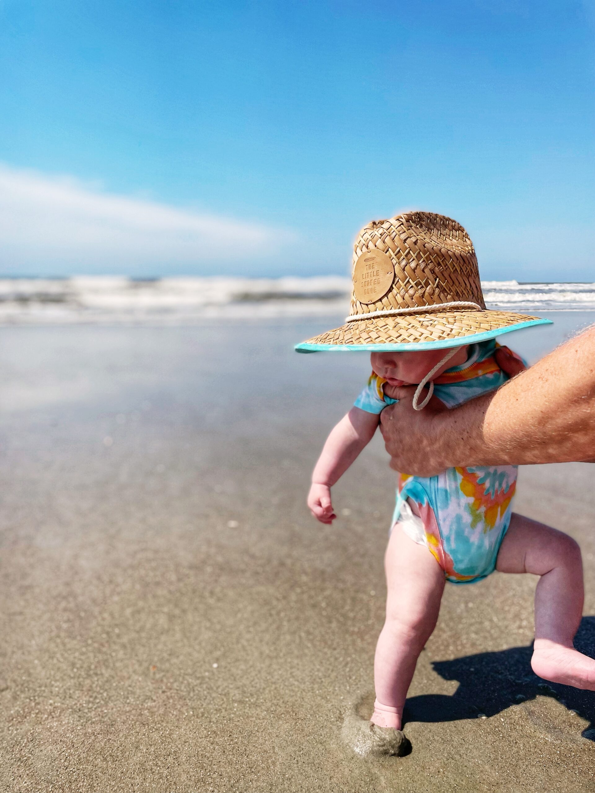 A baby wearing a large straw hat and a colorful onesie is held up by an adult hand while standing on a sandy beach near the ocean, with blue sky overhead.