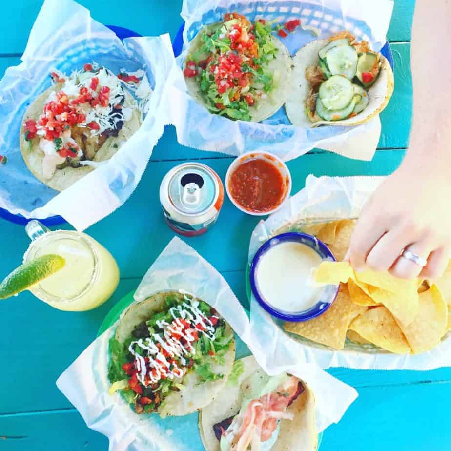 Colorful tacos with various toppings, tortilla chips with dip, salsa, a soda can, and a drink with a lime wedge on a bright turquoise table. A hand is reaching for a chip.