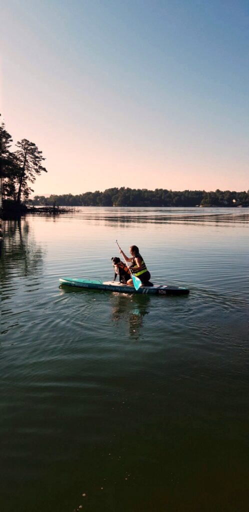 A person in a yellow life vest paddles a green kayak with a black and white dog sitting at the front on a calm lake, surrounded by trees under a clear sky.