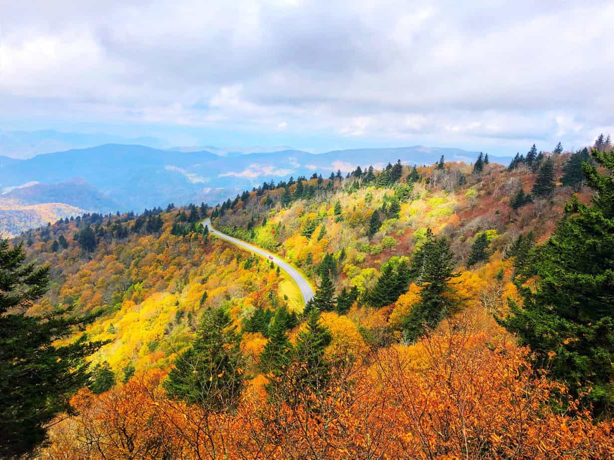 A winding road cuts through a forested mountain landscape, with vibrant autumn foliage in shades of orange, yellow, and green under a partly cloudy sky.