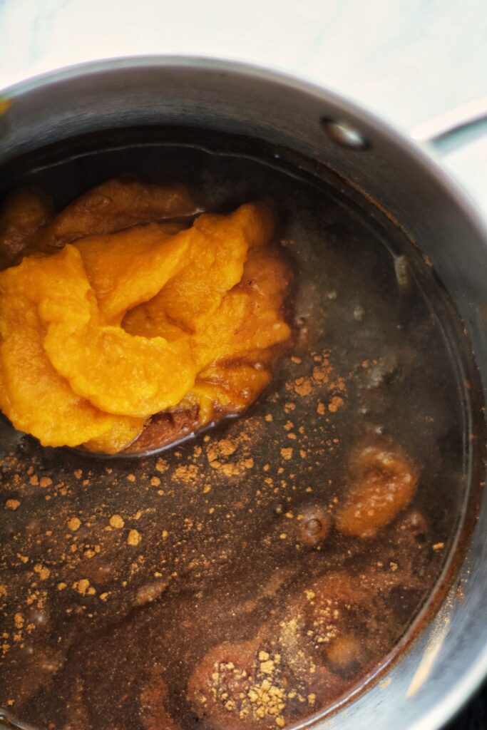 A close-up of a pot containing a dark liquid with spices and a mound of orange pumpkin puree, ready to be mixed together for cooking. Steam rises from the surface.