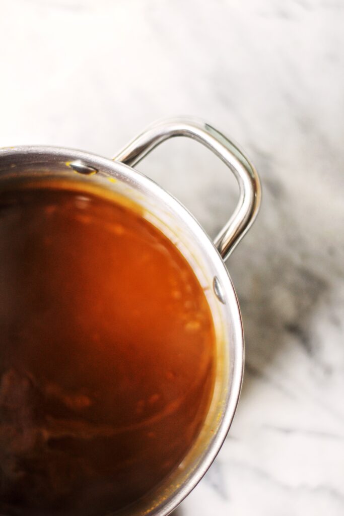 A stainless steel pot filled with brown liquid, possibly soup or sauce, sits on a white marble surface. Only half of the pot is visible, showing one handle and part of the contents.