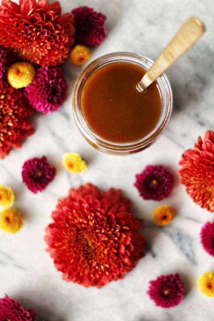 A jar of caramel sauce with a spoon in it sits on a marble surface, surrounded by red, pink, and yellow flowers.