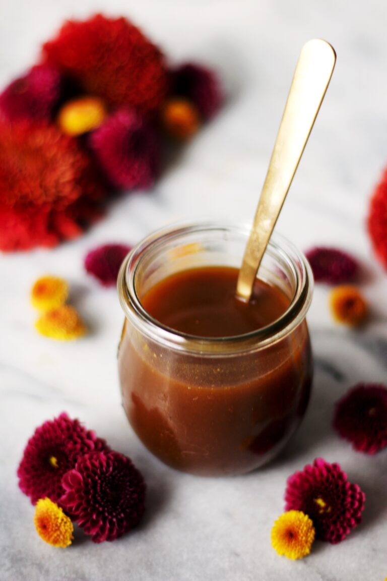 A glass jar filled with caramel sauce and a gold spoon, surrounded by red and yellow flowers on a marble surface.