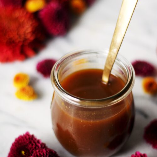 A small glass jar filled with pumpkin spice syrup and a gold spoon, surrounded by red and yellow chrysanthemum flowers on a white marble surface.