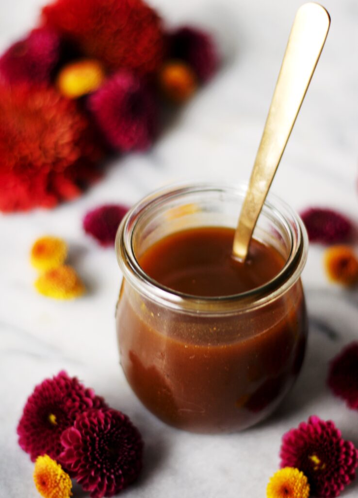 A small glass jar filled with pumpkin spice syrup and a gold spoon, surrounded by red and yellow chrysanthemum flowers on a white marble surface.