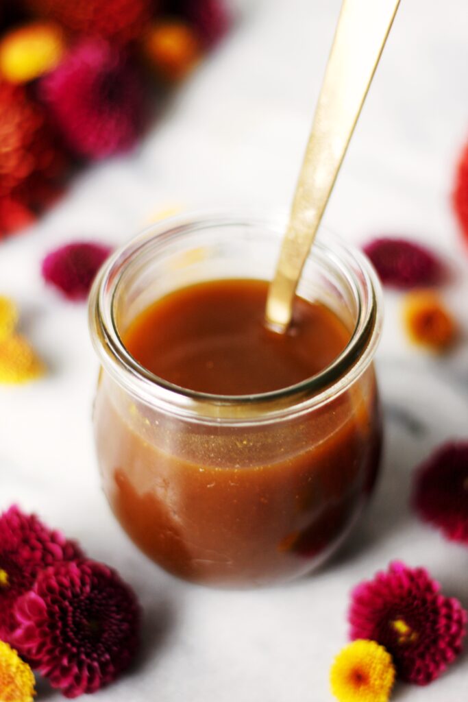 A small glass jar filled with caramel sauce, with a gold spoon inside, surrounded by purple and yellow flowers on a white surface.