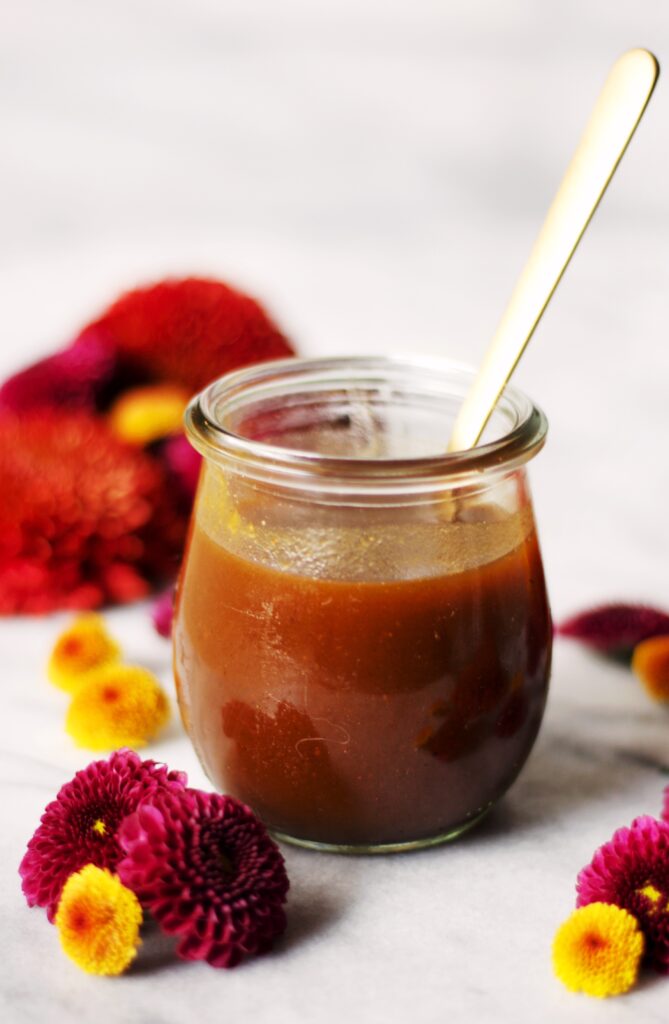 A glass jar filled with brown sauce or syrup, with a gold spoon inside. The jar is surrounded by red and yellow flowers, against a light, blurred background.