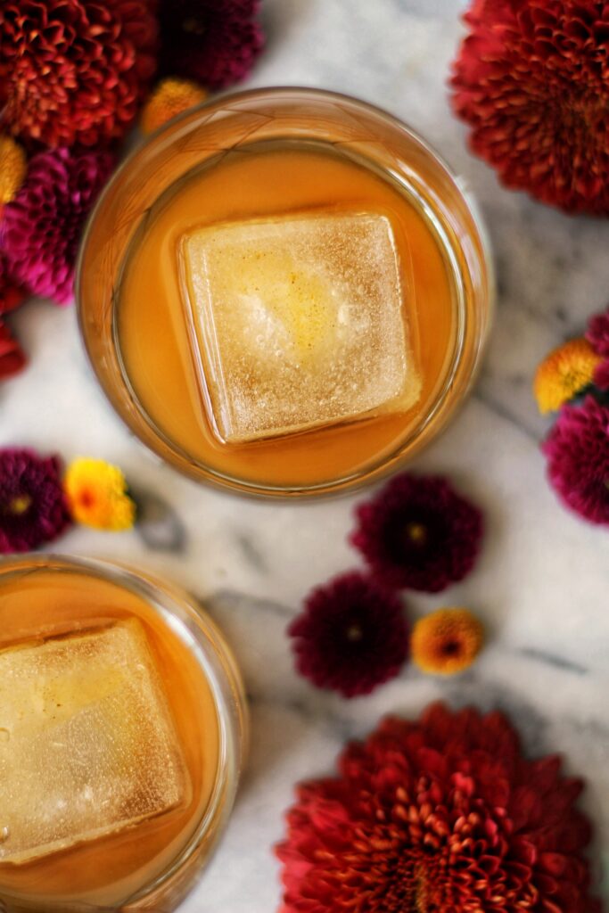 Two glasses of amber-colored drinks with large square ice cubes, surrounded by red and orange flowers on a marble surface.